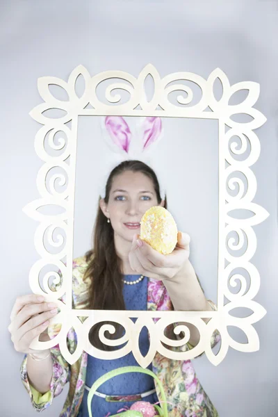 Pretty young woman wearing colorful jacket, blue dress and rabbit ears and holding yellow sequin Easter egg