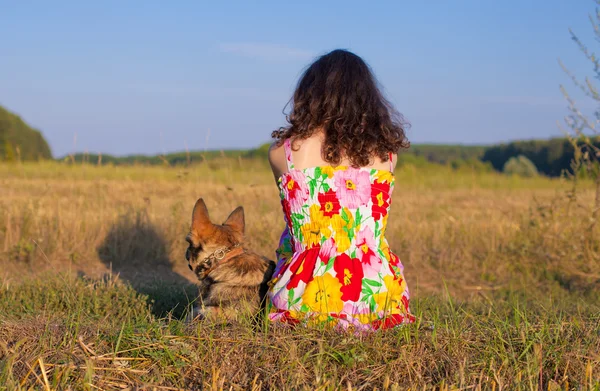 Young woman sitting with her dog back to camera on the meadow at sunset.