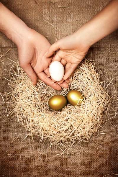 Men and women hands putting white egg to nest with golden eggs