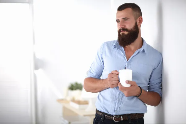 Young man standing near wall and holding cup of coffee in office