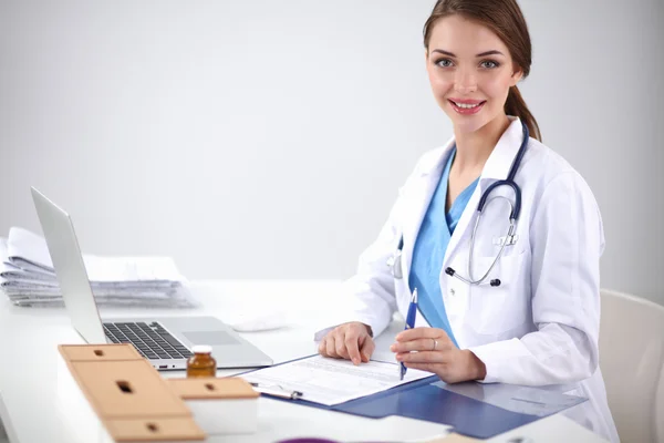 Beautiful young smiling female doctor sitting at the desk and writing.