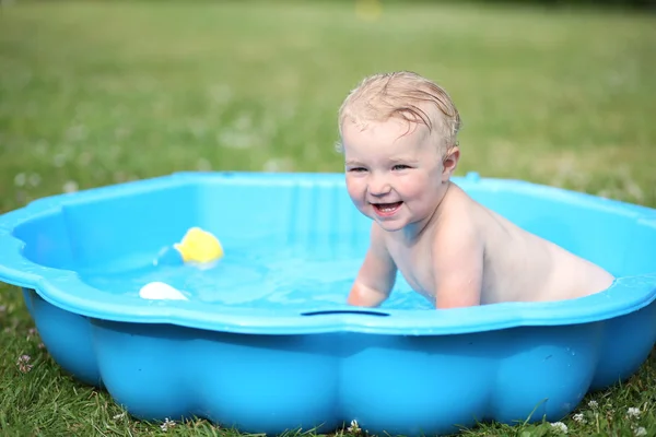 Pretty blonde baby girl playing with water in little plastic bath outdoors in the garden at the backyard of the house