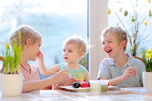 Group of cute children from one family, two twin brothers and their little toddler sister, decorating and painting Easter eggs sitting together in the kitchen on a sunny day. Selective focus on girl.
