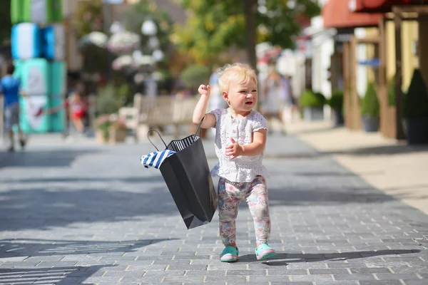 Cute baby girl standing or walking in the middle of the street in outlet village during sales with black shopping bag in her hands, crowd of people in the background