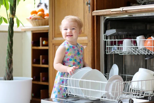Smiling blonde toddler girl helping in the kitchen taking plates out of dish washing machine