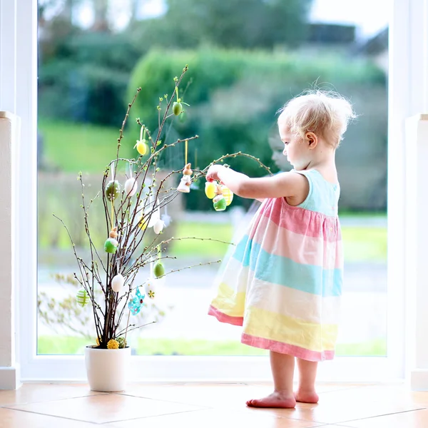 Cheerful blonde little toddler girl decorating cherry tree branches with Easter eggs standing indoors next to a big window with street view