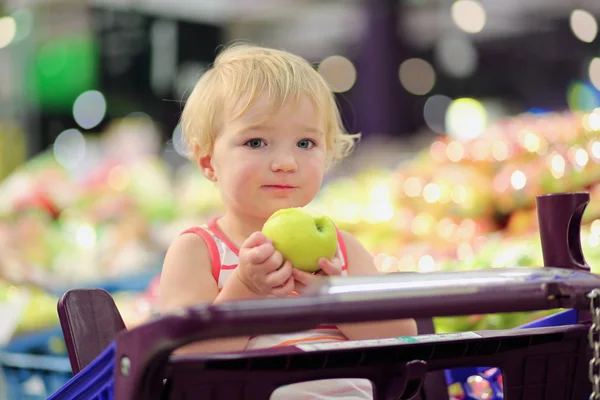 Funny little girl eating apple sitting in supermarket shopping cart