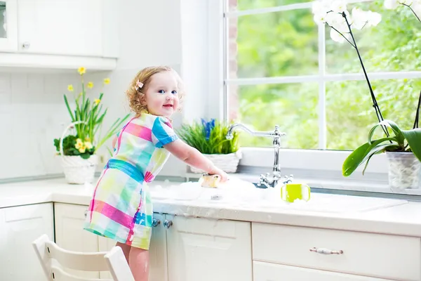 Cute curly toddler girl in a colorful dress washing dishes