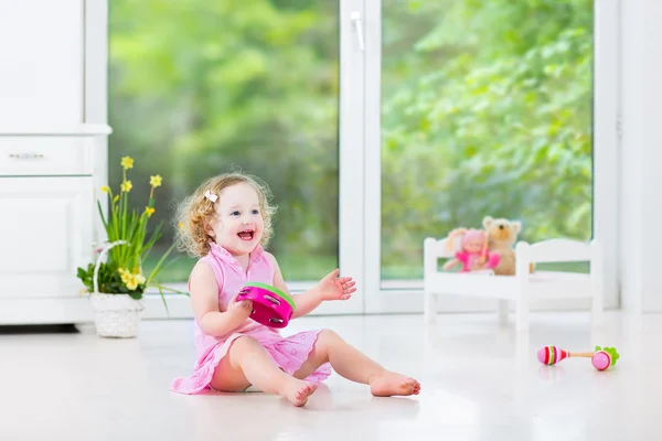 Cute curly toddler girl playing tambourine in a sunny white room