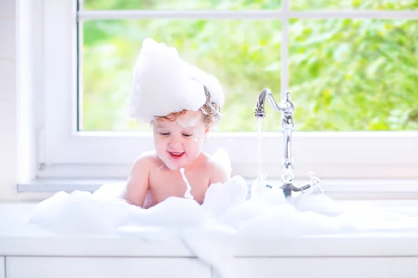 Funny baby girl playing with water and foam in a big kitchen sin