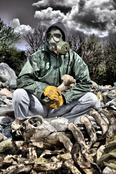 Man in a gas mask sitting on the garbage and holding a bone