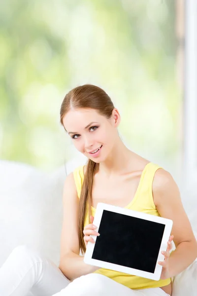 Beautiful girl sitting on a sofa against the window and shows the electronic tablet