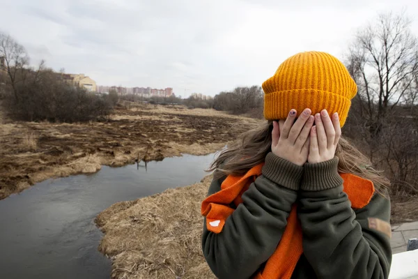 Sad teenager in orange knitten hat and scarf closed her face by hands, stand alone near the scorched field. Spring time