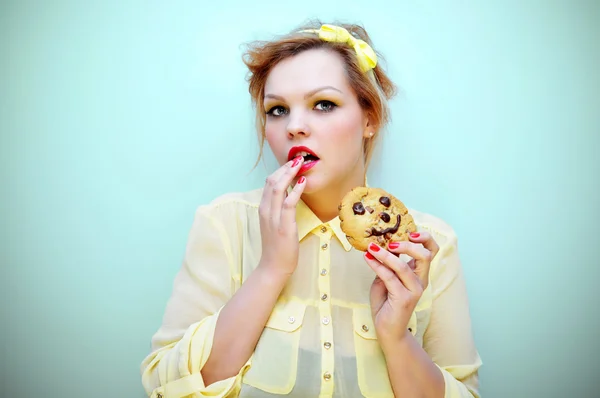 Young attractive woman with red hair and yellow bow headband wearing red lipstick and a yellow chiffon blouse is eating a smiling chocolate chip cookie.