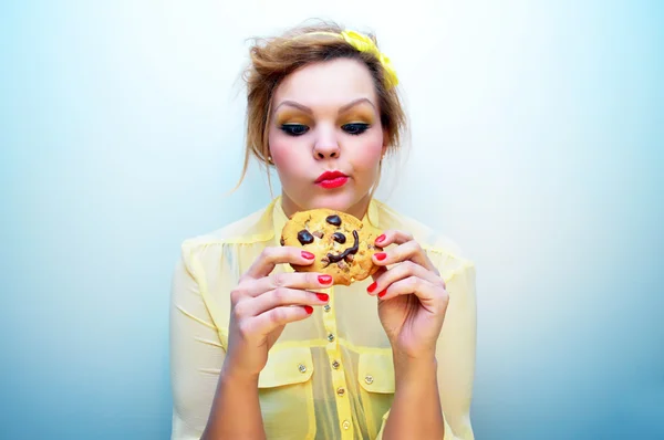 Young attractive woman with red hair and a yellow bow headband wearing a yellow chiffon blouse is holding a smiling chocolate chip cookie and looking at it with an indecisive look.
