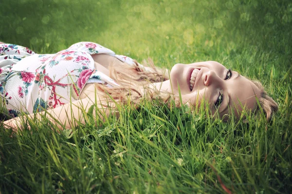 Beautiful smiling young woman wearing a flower print summer dress lying on green grass on the meadow.