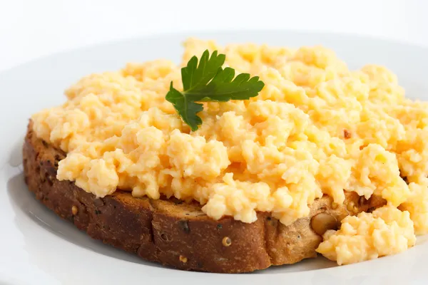 Soft scrambled eggs with parsely leaf on brown toast on white plate.