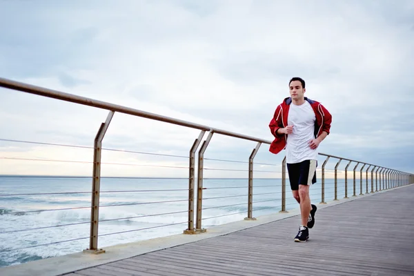Young athlete man in white t-shirt and sports a red windbreaker runs beach pier
