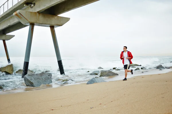 Young athlete man in white t-shirt and sports a red windbreaker runs along the beach enjoying the beautiful sea