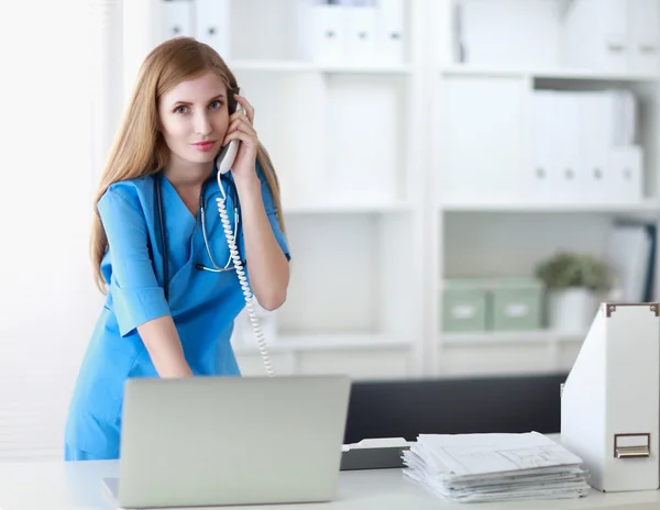 Young woman doctor in white coat at computer using phone