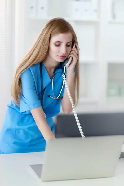 Young woman doctor in white coat at computer using phone