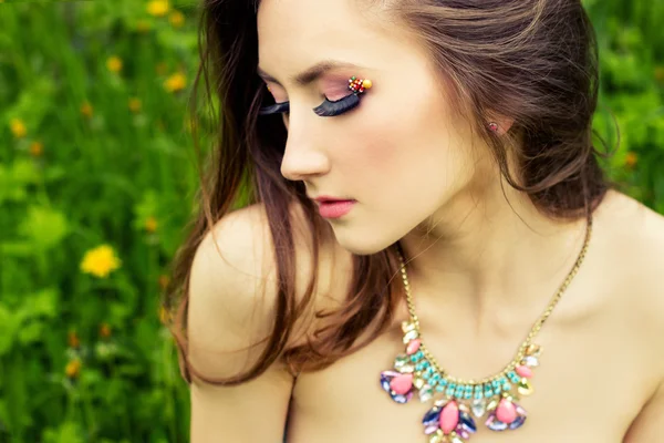 Beautiful young girl with long hair and beautiful makeup with necklace on the neck sitting on the grass in the garden