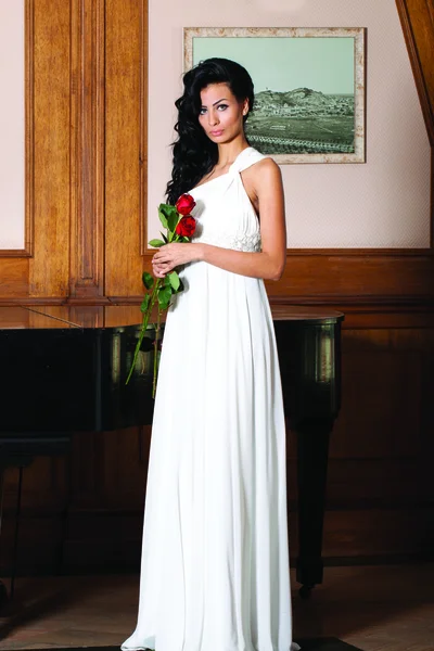 Full length indoor portrait of sexy bride with black long hair. Holding red roses.