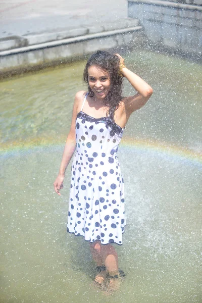 Woman in a wet dress and wet brown hair posing on the background of the rainbow from the fountain