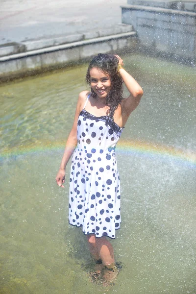 Woman in a wet dress and wet brown hair posing on the background of the rainbow from the fountain