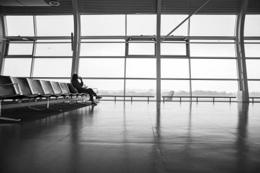 Havaalanı terminalindeki büyük vitraylı pencerenin önünde boş bir sıra koltukta oturan bir kız uçuş için bekliyor..