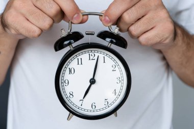 Beyaz tişörtlü bir adam elinde zili olan şık siyah bir çalar saat tutuyor. Alarm saatinde, sekizincinin başında. Kalkma zamanı.