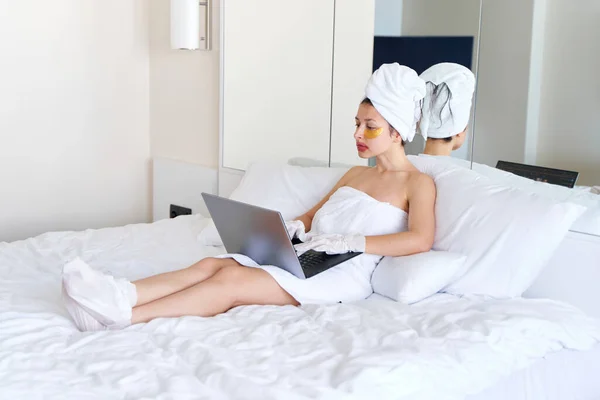 After a shower, a girl wrapped in a towel works on a laptop and uses cosmetic patches for the skin under the eyes, lips and gloves to moisturize her hands and feet. Cosmetic trends for body care at home.