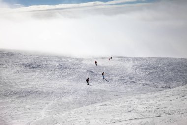 Kayakçılar ve snowboardcular kar yamacında yokuş aşağı kayıyorlar ve kış sabahı sisli bir havada güneşleniyorlar. Kafkasya Dağları, Gürcistan, Gudauri bölgesi.