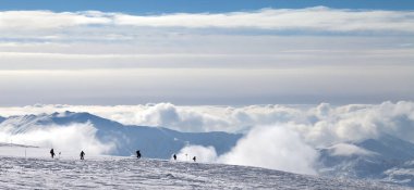 Kar yamacında kayakçıların ve snowboardcuların siluetlerinin panoramik görüntüsü ve kış akşamları güzel bulutlarda dağların görünmesi. Kafkasya Dağları, Gürcistan, Gudauri bölgesi.