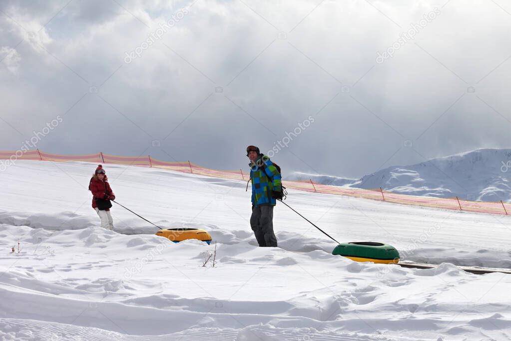 Father and daughter with snow tube in snowy sunlight mountains with gray cloudy sky. Caucasus Mountains, Georgia, region Gudauri at winter.