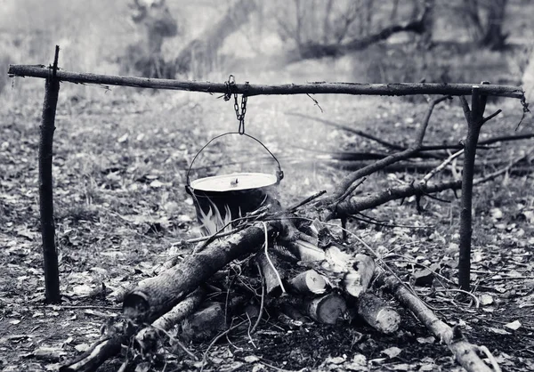 Cooking in old sooty cauldron on campfire at forest. Black and white retro toned image.