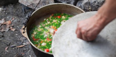 Kamp ateşinde eski, ıslak kazanda taze pişmiş sıcak çorba ve ot kılıflı bir erkek eli. Açık hava kamp yemekleri. Seçici odak.