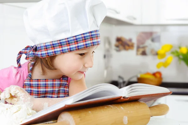 Little baker in hat preparing cookies with cookbook — Stock Photo, Image