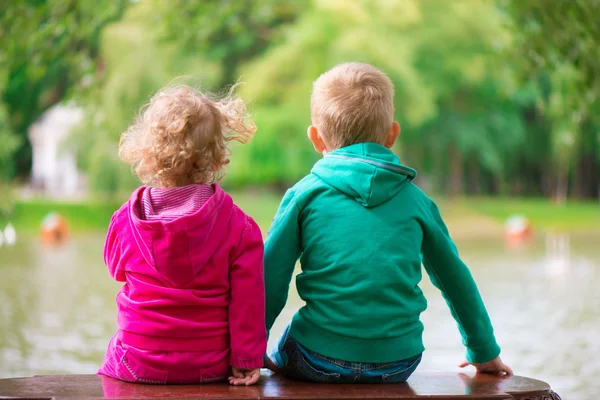 Little sister and brother sitting on bench at pond — Stock Photo, Image