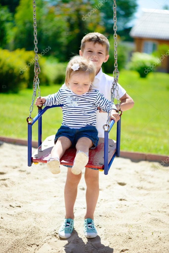 Little brother and sister swinging in summer park 