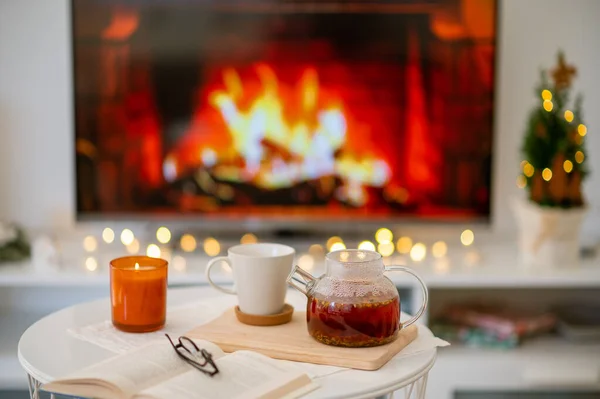 Photo of cozy place with tea cup, teapot, candle and book with glasses with fireplace imitation on tv on background
