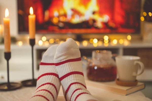 Close up cozy photo of feet in striped socks on side table with candles, teapot and cup  bevor fireplace