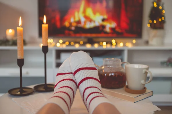 Close up cozy photo of feet in striped socks on side table with candles, teapot and cup  bevor fireplace imitation on TV