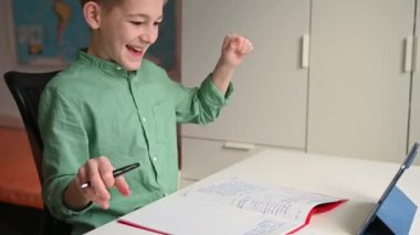 Online ders sırasında tablet kullanan ve deftere yazı yazan genç bir çocuk.