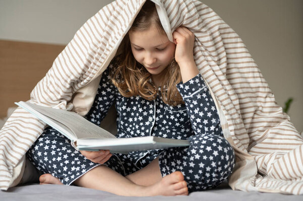 Милая маленькая девочка-подросток сидит и читает книгу на кровати под одеялом 