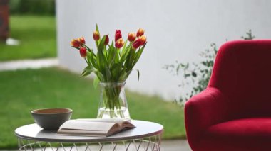 Kırmızı rahat sandalye, arka bahçedeki rahat modern terasta içinde kitap ve vazo olan küçük masa.