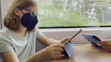 Pretty teen girl in medical mask playing with tablet in train during traveling
