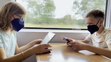 Two happy teen siblings children in medical masks playing with tablets in train during traveling 