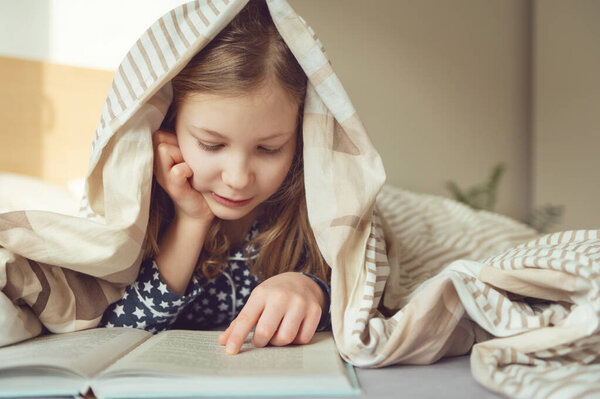 Милая маленькая девочка-подросток лежит и читает книгу на кровати под одеялом 