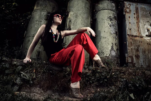 Chica en pantalones rojos y gafas sentado sobre fondo industrial —  Fotos de Stock